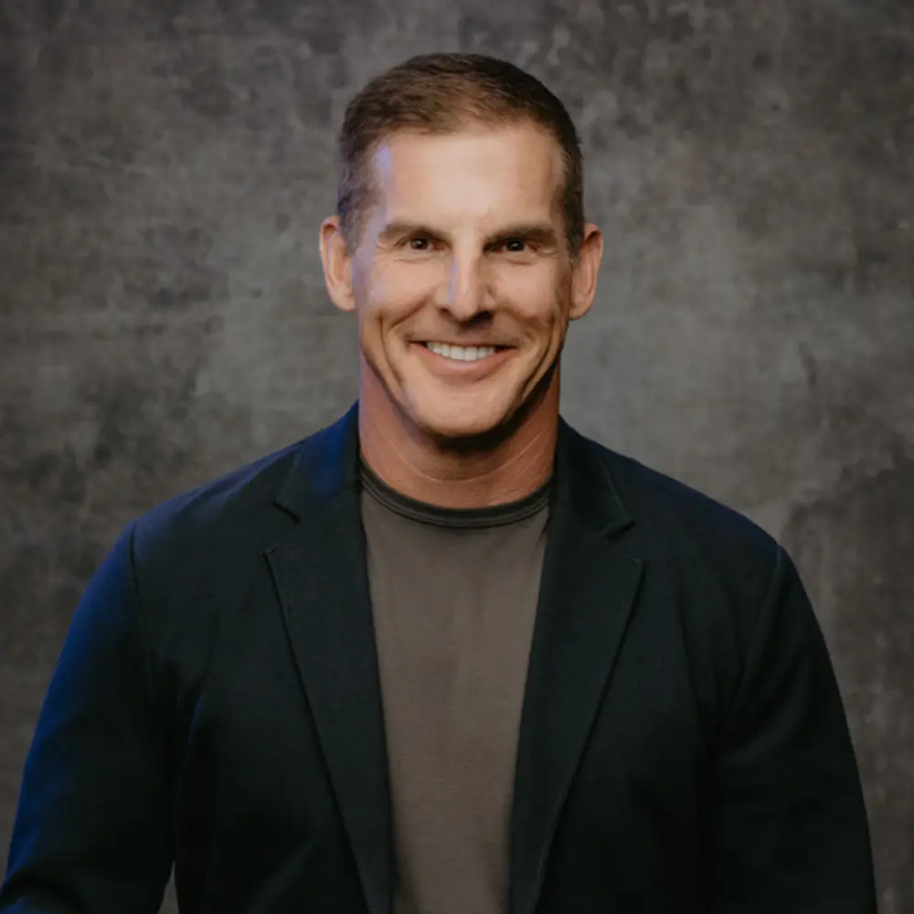A smiling man in a black blazer and dark shirt against a gray backdrop.