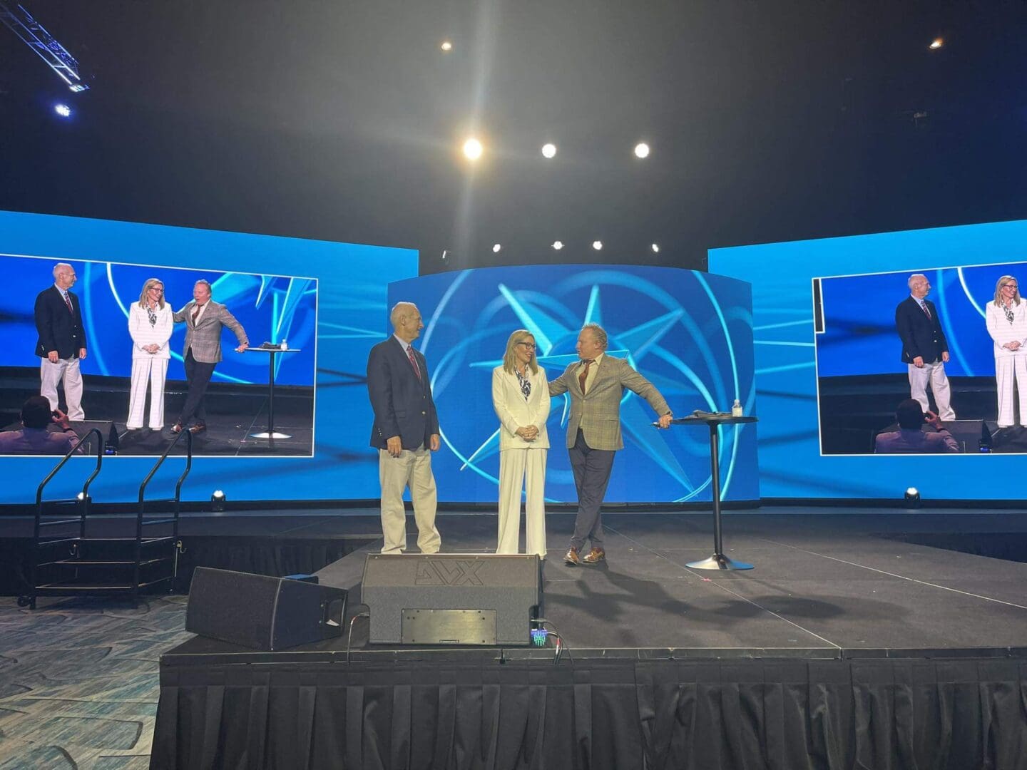 Three people on stage with blue screens and spotlights.