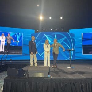 Three people on stage with bright blue screens behind them.